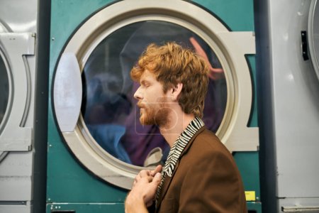 Foto de Vista lateral del joven pelirrojo de moda con chaqueta posando cerca de la lavadora en la lavandería de monedas - Imagen libre de derechos