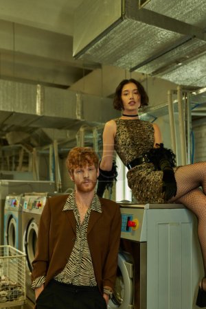 Photo for Fashionable young multiethnic couple in trendy outfits posing and looking at camera in coin laundry - Royalty Free Image