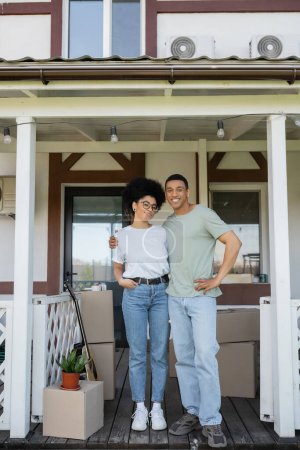 sonriente afroamericano hombre abrazando novia y mirando a la cámara en el porche de nueva casa