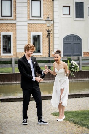 Photo for Groom opening champagne bottle near joyful african american bride on street, outdoor celebration - Royalty Free Image