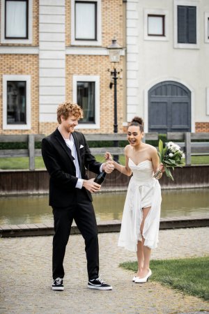 Photo for Wedding in city, groom opening champagne near excited african american bride with flowers on street - Royalty Free Image