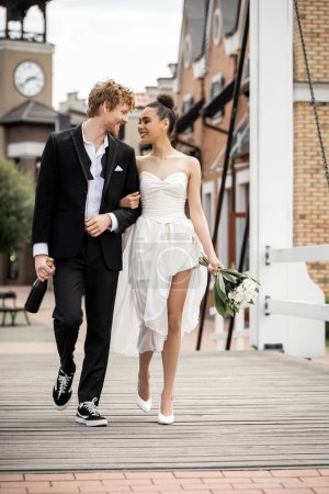 Photo for Wedding outdoors, multiethnic newlyweds with champagne and flowers walking on city bridge - Royalty Free Image