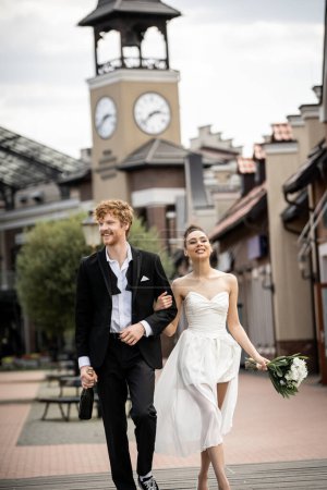 Photo for Romantic and cheerful interracial newlyweds with champagne and flowers walking on urban street - Royalty Free Image