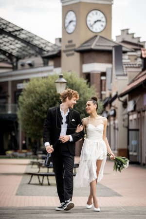 Hochzeit im Freien, junges multiethnisches Paar in eleganter Kleidung mit Champagner und Blumen auf der Straße