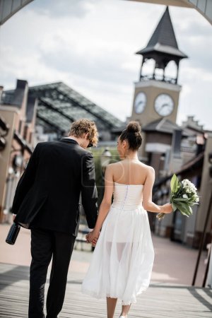 Hochzeit in moderner Stadt, Rückansicht eines eleganten multiethnischen Paares mit Champagner und Blumen auf der Straße