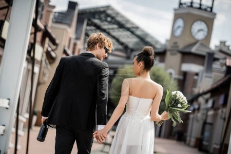 Rückansicht eines eleganten gemischtrassigen Paares mit Champagner und Blumen in der Stadt, Hochzeit im Freien