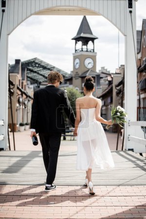 Photo for Back view of romantic multiethnic couple holding hands and walking on bridge, outdoors wedding - Royalty Free Image