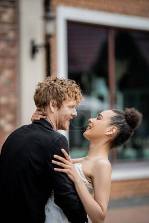 Photo for Wedding in european city, side view of carefree and elegant multiethnic couple embracing on street - Royalty Free Image