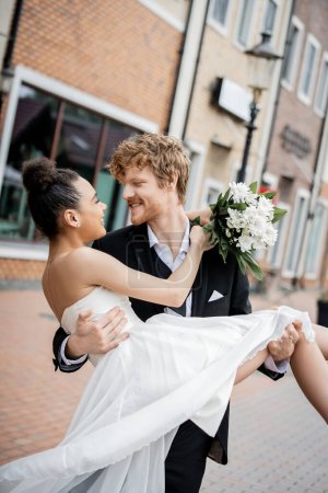 Photo for Urban romance, redhead groom holding african american bride with wedding bouquet on street - Royalty Free Image