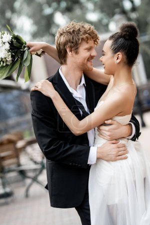 interracial newlyweds in elegant attire embracing on street, outdoors wedding, banner