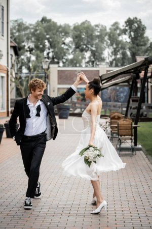 boda en la ciudad moderna, elegante pareja multiétnica cogida de la mano y bailando en la calle, felicidad