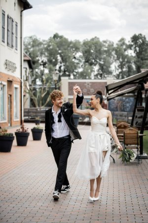 overjoyed multiethnic newlyweds dancing and holding hands on city street, outdoor celebration