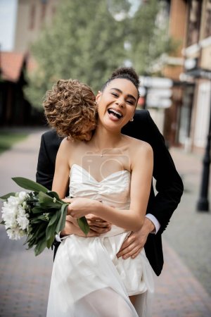 marié rousse embrassant mariée afro-américaine riante avec bouquet, célébration de mariage en ville