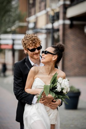 Photo for Interracial couple with burgers and orange juice near city fountain, wedding attire, sunglasses - Royalty Free Image