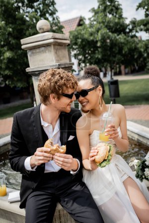 multiethnic newlyweds celebrating wedding near city fountain, burgers, orange juice, snack