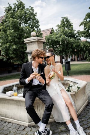 happy interracial couple in wedding attire and sunglasses with burgers and juice near city fountain