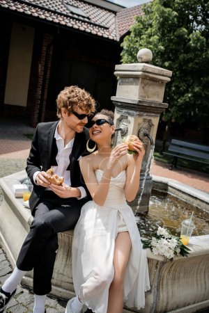 Photo for Multiethnic newlyweds in sunglasses snack with burgers and orange juice near city fountain - Royalty Free Image
