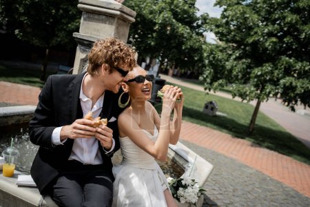 Photo for Excited multiethnic couple celebrating wedding near city fountain, sunglasses, burgers, orange juice - Royalty Free Image