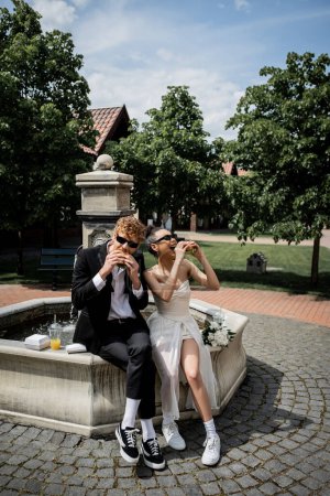 elegant interracial newlyweds in sunglasses eating burgers near fountain in european city