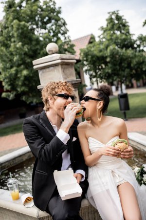 interracial couple in wedding attire and sunglasses eating burger together near fountain in city