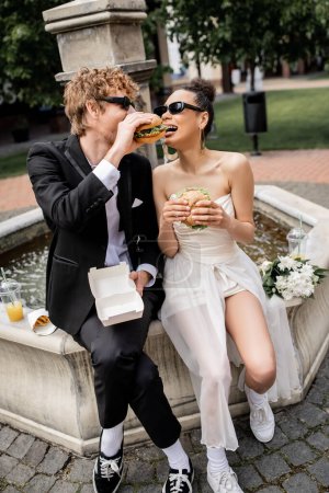 elegant interracial newlyweds in sunglasses eating burger together, city street, fountain, fun