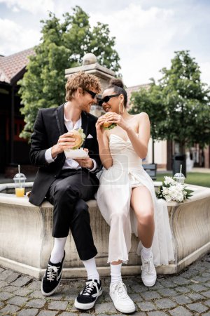 joyful interracial newlyweds in sunglasses holding burgers and looking at each other near fountain