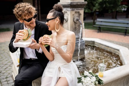 outdoor wedding, interracial couple in sunglasses snacking with burgers at fountain in european city