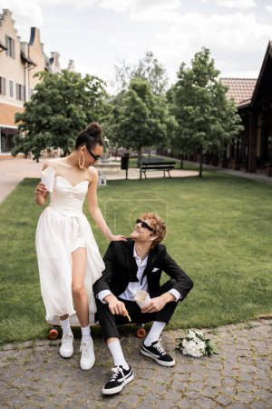 Photo for Redhead groom sitting on longboard near african american bride with french fries, outdoor wedding - Royalty Free Image