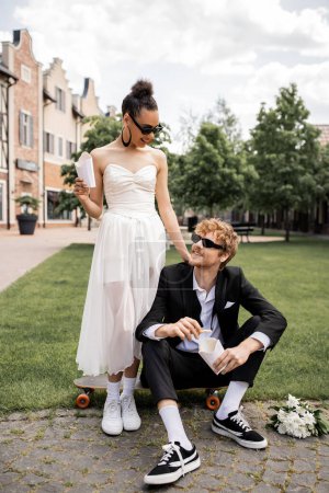 Photo for Elegant african american bride near groom sitting on longboard on street, french fries, sunglasses, - Royalty Free Image