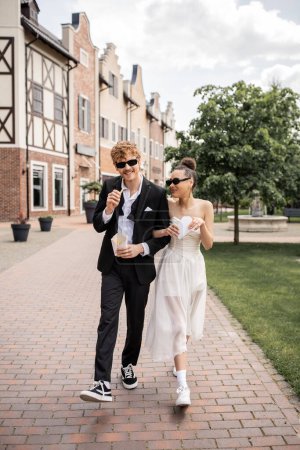 Photo for Redhead man and african american woman walking with french fries in city, wedding attire, sunglasses - Royalty Free Image