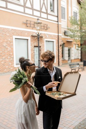 stylish multiethnic newlyweds with flowers in pizza on urban street, wedding attire, sunglasses