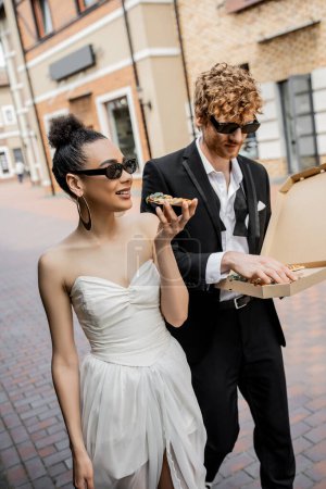 outdoor celebration, interracial couple walking with pizza in city, wedding attire, sunglasses