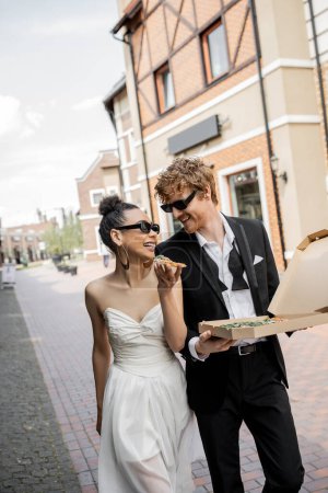 Photo for Love in city, happy multiethnic newlyweds in sunglasses walking with pizza on street - Royalty Free Image