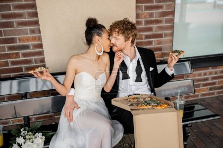 moderne gemischtrassige Brautpaare mit Pizza auf Bank in der Stadt, Feiern im Freien, Glück