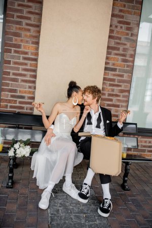 elegant multiracial couple with pizza kissing on bench, wedding in city, outdoor celebration