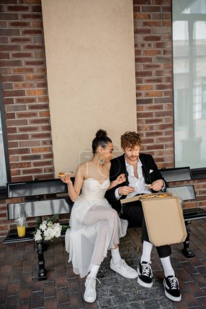 multiethnic couple in wedding attire sitting on bench with pizza near bouquet, outdoor celebration