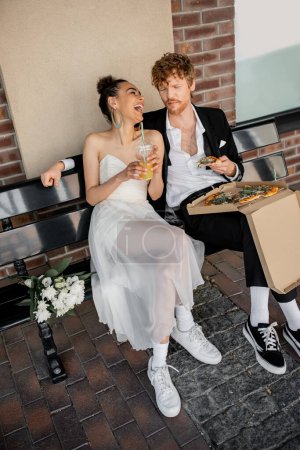 african american bride with orange juice laughing near redhead groom and pizza on bench in city