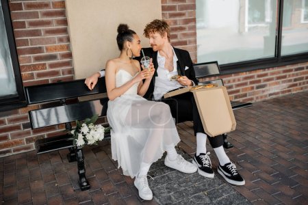 glückliches gemischtrassiges Brautpaar mit Pizza und Orangensaft, die sich auf der Bank anschauen, Hochzeit in der Stadt
