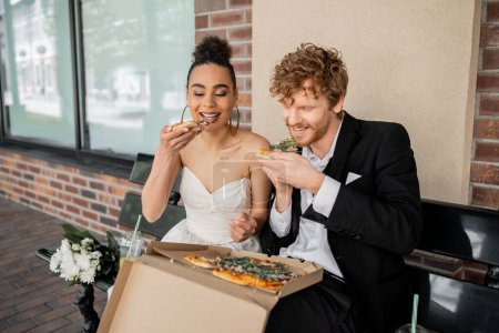 Photo for Smiling african american bride and redhead groom eating pizza on bench, wedding celebration in city - Royalty Free Image