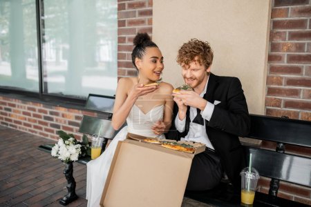 cheerful, elegant multiethnic newlyweds eating pizza near flowers and orange juice on bench in city