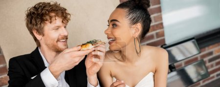 happy redhead man feeding elegant african american bride with pizza, urban setting, banner