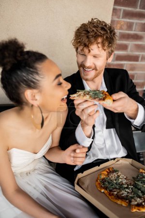 rothaariger Bräutigam füttert überglückliche Braut mit leckerer Pizza auf Bank, Outdoor-Hochzeit in der europäischen Stadt
