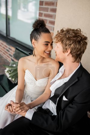 cheerful interracial couple holding hands and looking at each other on bench, wedding in city