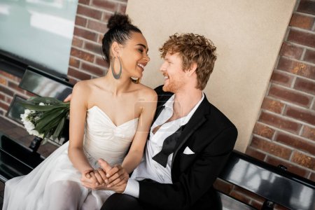 joven pareja multiétnica sonriendo el uno al otro y tomados de la mano en el banco, boda, entorno urbano
