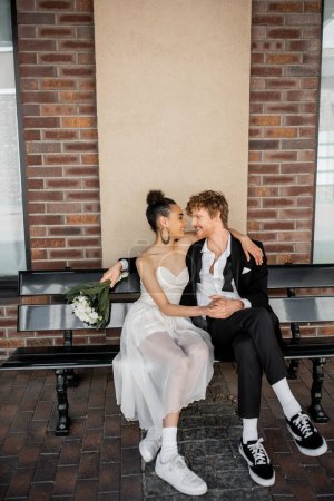 mariage en ville, heureux marié rousse avec bouquet assis face à face avec la mariée afro-américaine