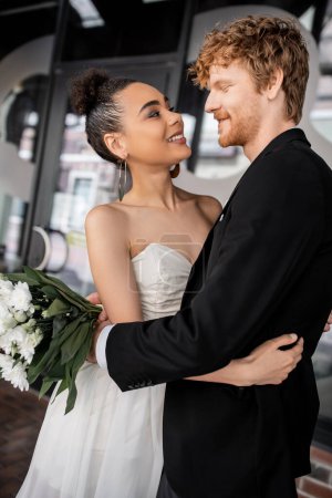 Photo for Overjoyed and elegant interracial couple in wedding attire embracing on city street - Royalty Free Image
