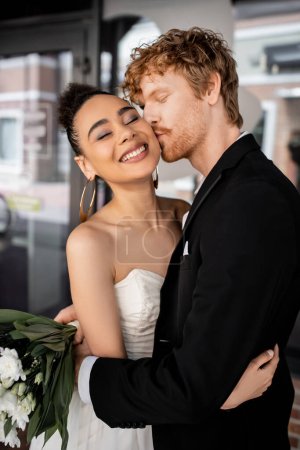 redhead groom in black suit embracing and kissing stylish african american bride on city street