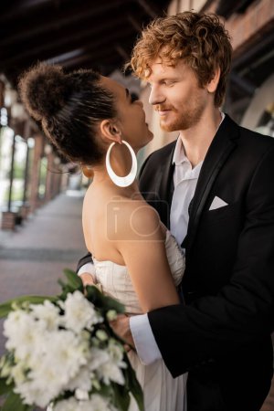 Photo for Joyful interracial couple embracing and looking at each other, wedding attire, urban setting - Royalty Free Image