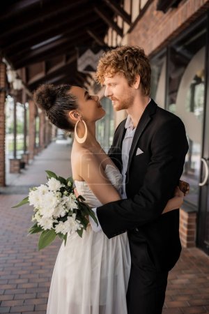 Photo for Side view of stylish multiethnic man with flowers looking at african american bride on city street - Royalty Free Image