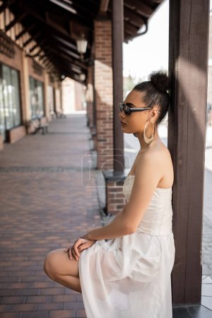 Photo for Side view of charming african american woman in wedding dress and sunglasses on urban street - Royalty Free Image